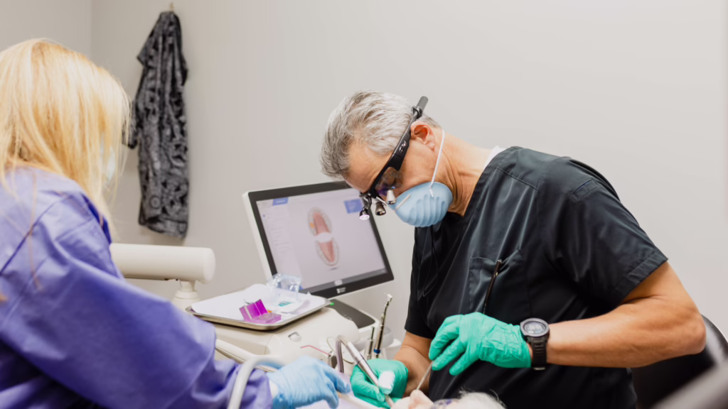 Dentist and assistant performing a dental procedure on a patient; computer screen shows a tooth diagram.